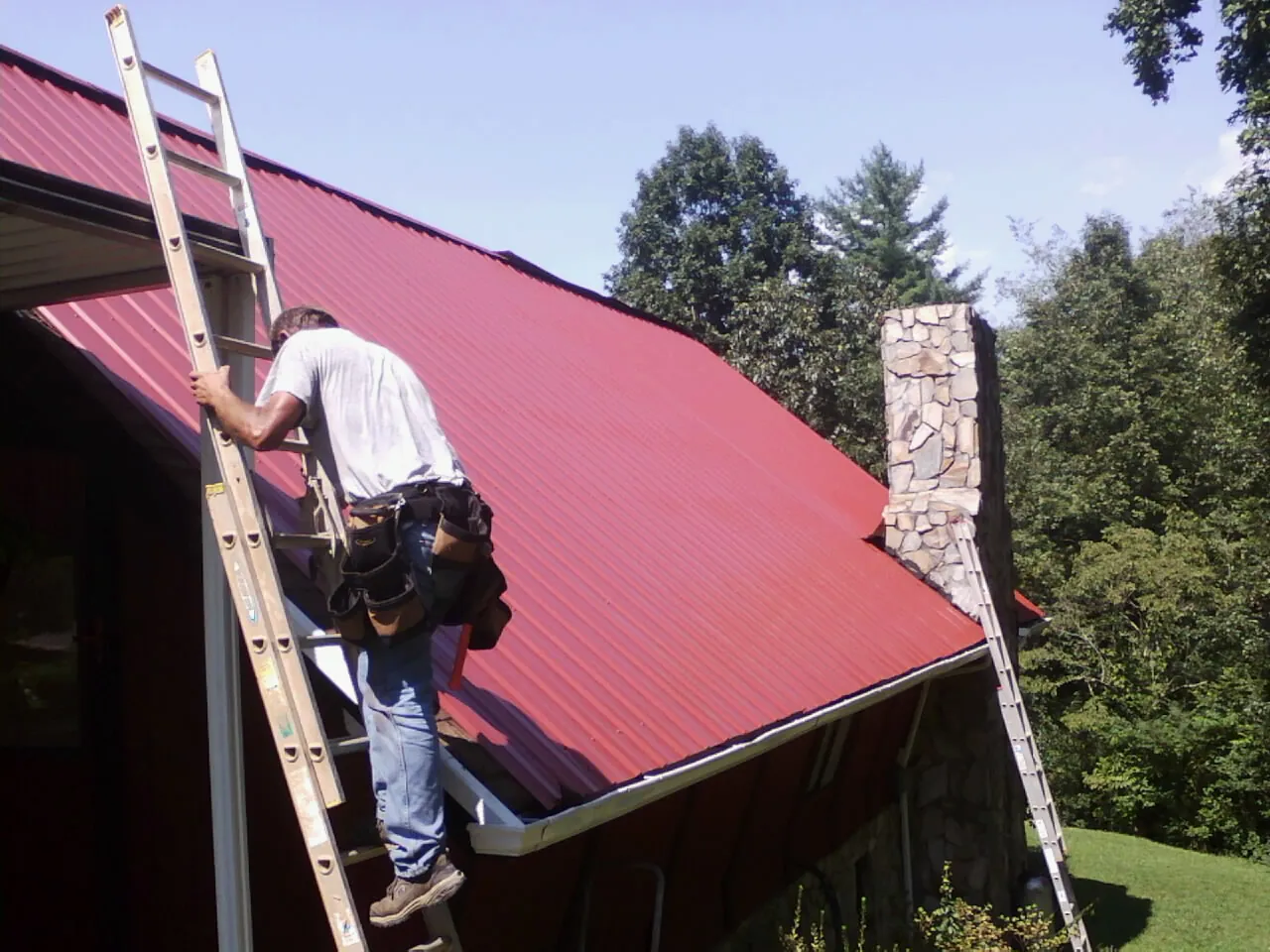 Roofer on a ladder beside a red metal roof and stone chimney — Soffit & Fascia Repair by Roofing Brothers in Traphill